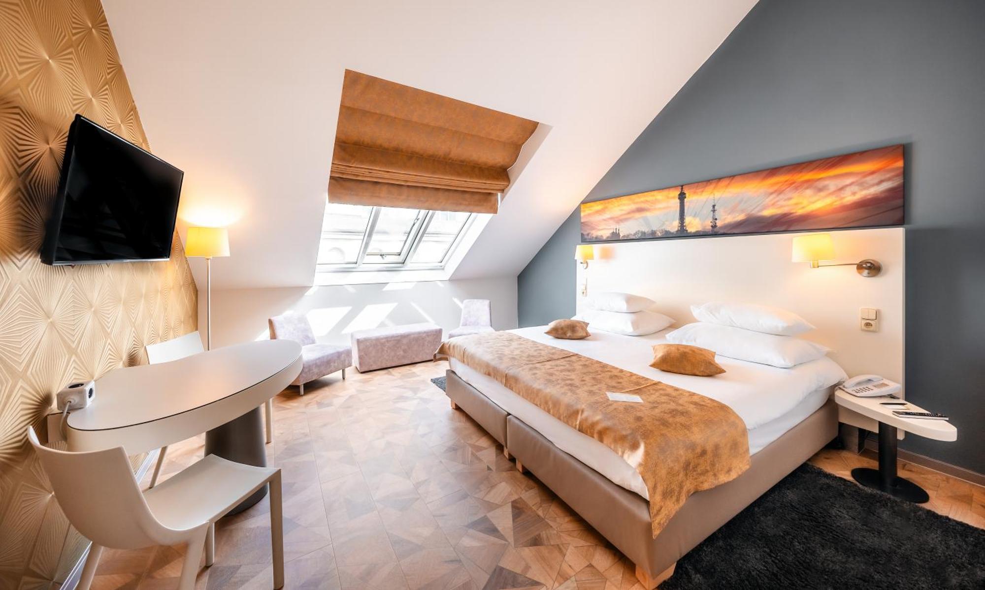 Grandium Hotel Prag Eksteriør billede The photo shows a stylish and modern hotel room with an angled ceiling. It features a large bed with white linens and decorative pillows at the center. A soft, fur-like throw covers part of the bed. There are two bedside lamps and a wall-mounted TV o