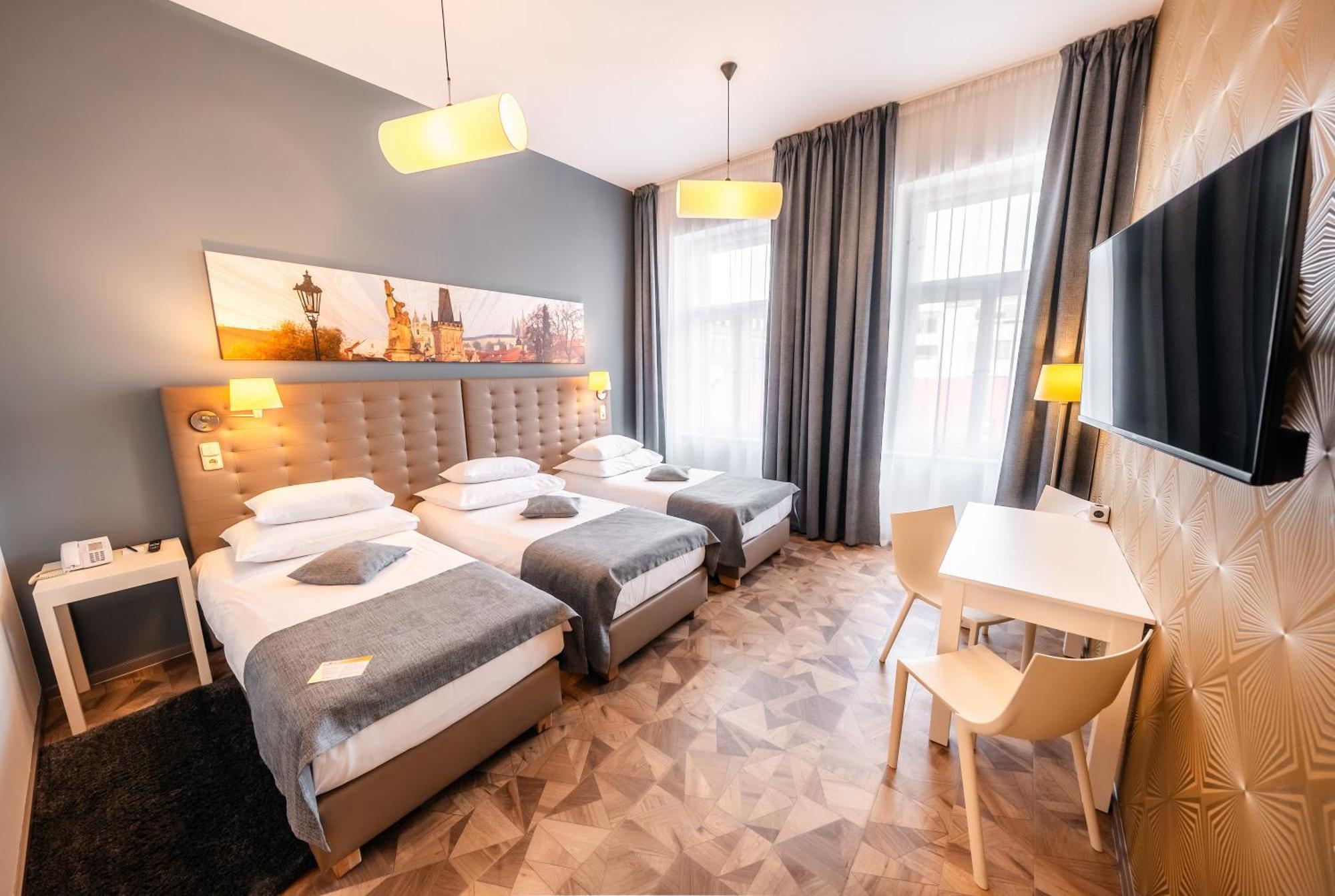 Grandium Hotel Prag Eksteriør billede The photo shows a modern hotel room featuring three single beds, each with white bedding and gray throws. The walls are painted gray, and artwork depicting a cityscape is displayed above the headboard. There are two stylish pendant lights hanging fro