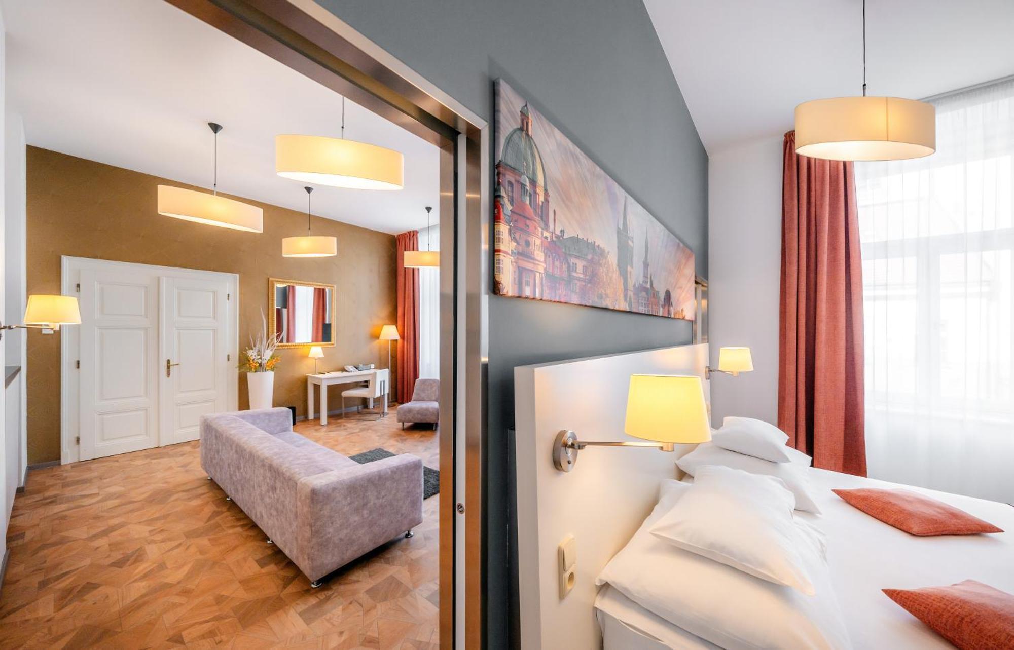 Grandium Hotel Prag Eksteriør billede The photo shows a stylish hotel room with modern decor. In the foreground, there is a bed with white pillows, and wall-mounted lamps provide soft lighting. Behind the bed, there's an open doorway leading to a living area featuring a light-colored sof