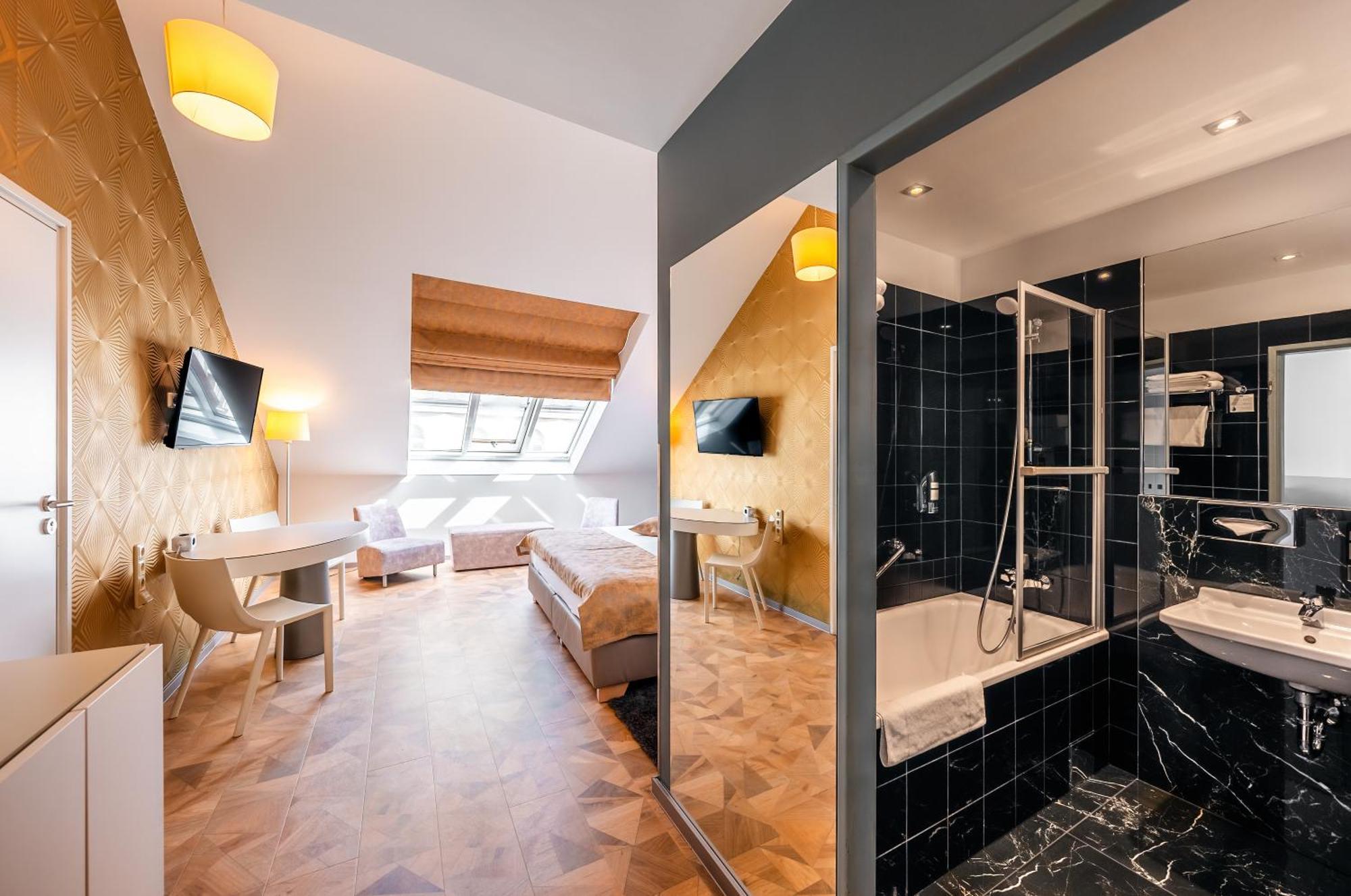Grandium Hotel Prag Eksteriør billede The photo shows an interior of a hotel room featuring a modern design. On one side, there are two beds with white linens, a desk, and a television mounted on the wall. The room has sloped ceilings and large windows that let in natural light. 

Adjace