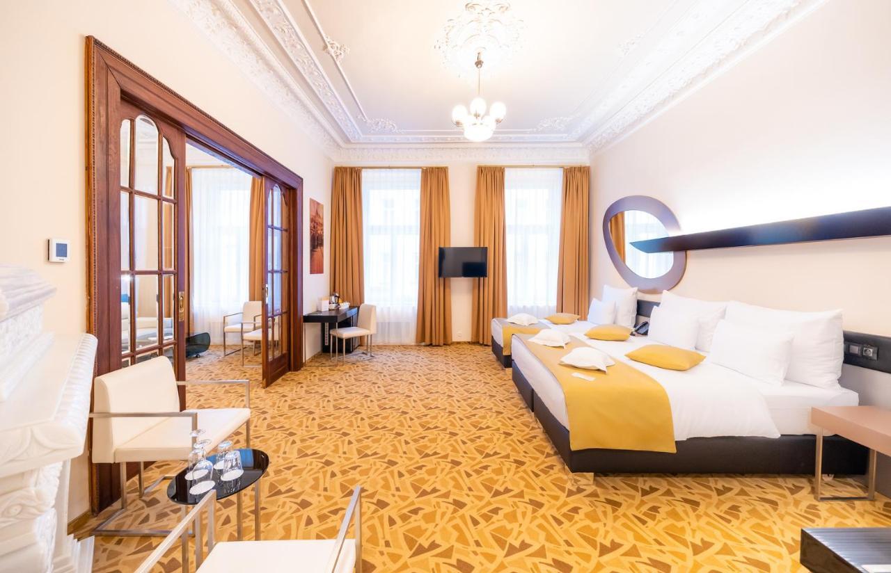 Grandium Hotel Prag Eksteriør billede The photo shows a spacious hotel room with an elegant design. There are two large beds with white bedding, accented with yellow pillows. The room has tall windows with flowing curtains that allow natural light to brighten the space. A flat-screen tel